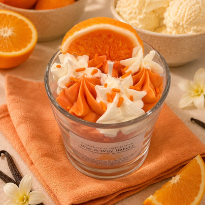 A clear glass jar candle filled with orange and white whipped frosting-like decoration and orange sprinkles, topped with an orange slice. The candle sits on an orange cloth with orange slices, vanilla beans, flowers, and bowls of ice cream in the background.