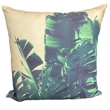 A square throw pillow featuring a tropical banana leaf pattern in shades of green and teal over a warm beige background, shown against a plain white studio backdrop.
