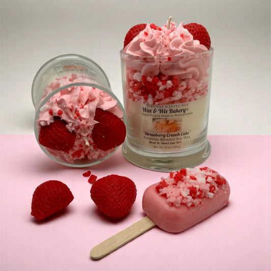 A strawberry-themed wax candle set displayed on a pink surface against a light gray background. The set includes a tall glass jar candle with pink whipped wax, red strawberries, and crushed candy cane pieces, a tipped-over glass jar showing the wax and ingredients, a pink popsicle-shaped wax piece on a wooden stick, and two loose strawberries.