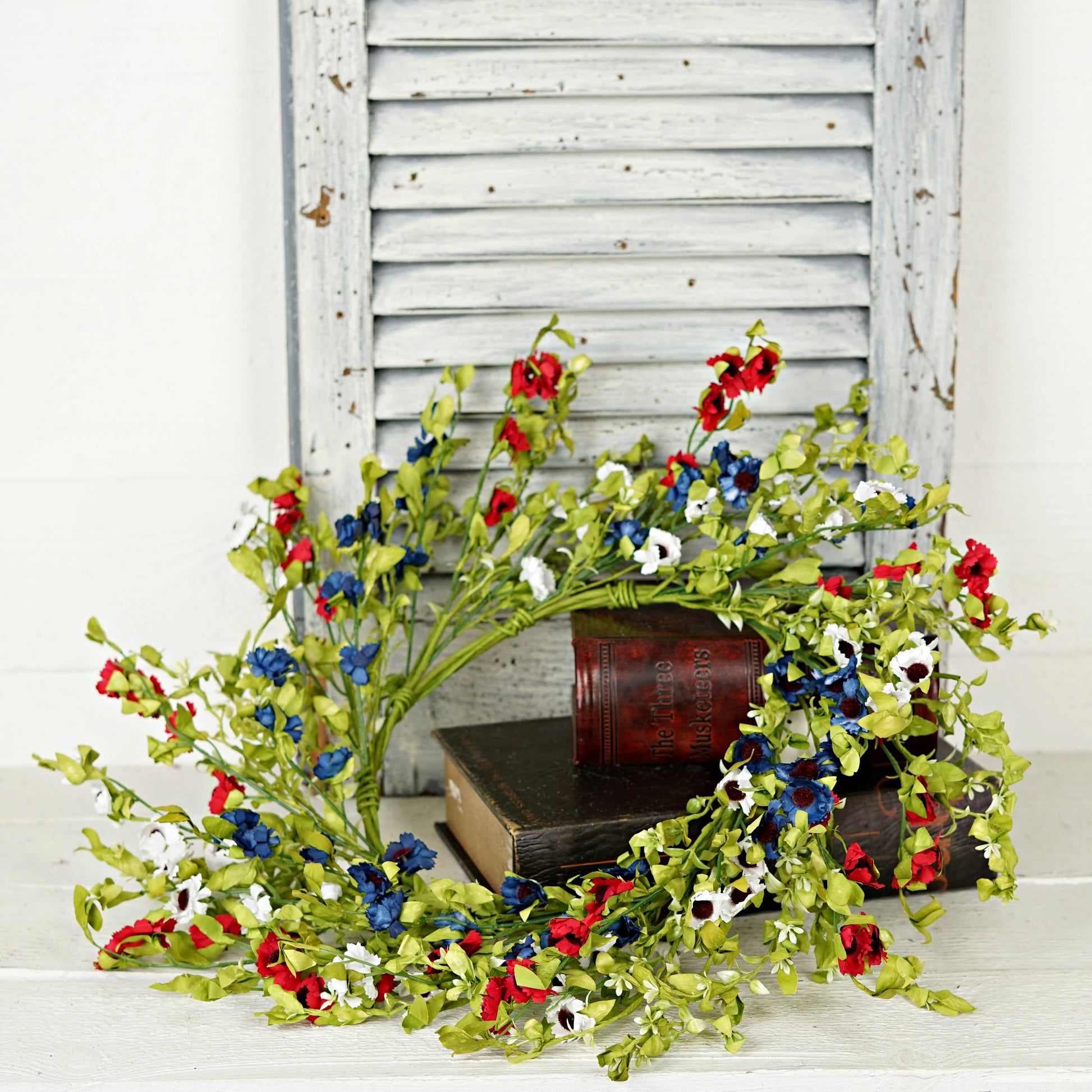 Decorative floral branch with red, blue, and white flowers on a wooden surface with a white shutter background.