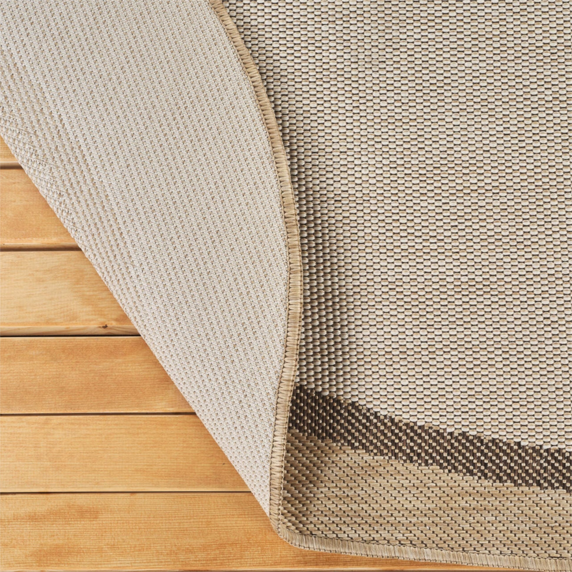 Close-up of a woven rug corner with one side folded back over a light wood floor, showing the rug's beige and brown textured weave and backing.