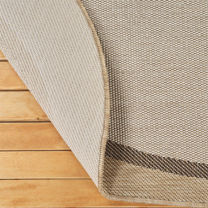 Close-up of a woven rug corner with one side folded back over a light wood floor, showing the rug's beige and brown textured weave and backing.