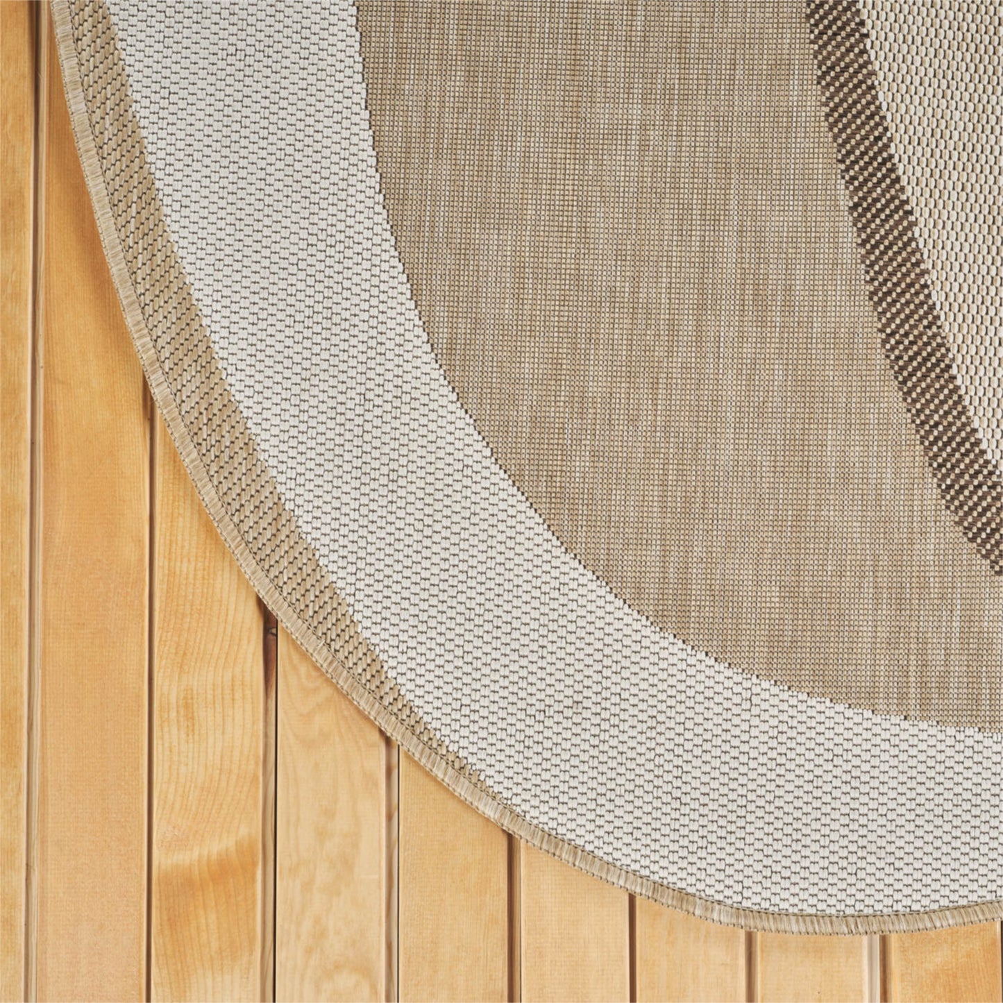 A close-up of a woven oval rug with a thick cream border and a tan center, laid over a light wood plank floor.