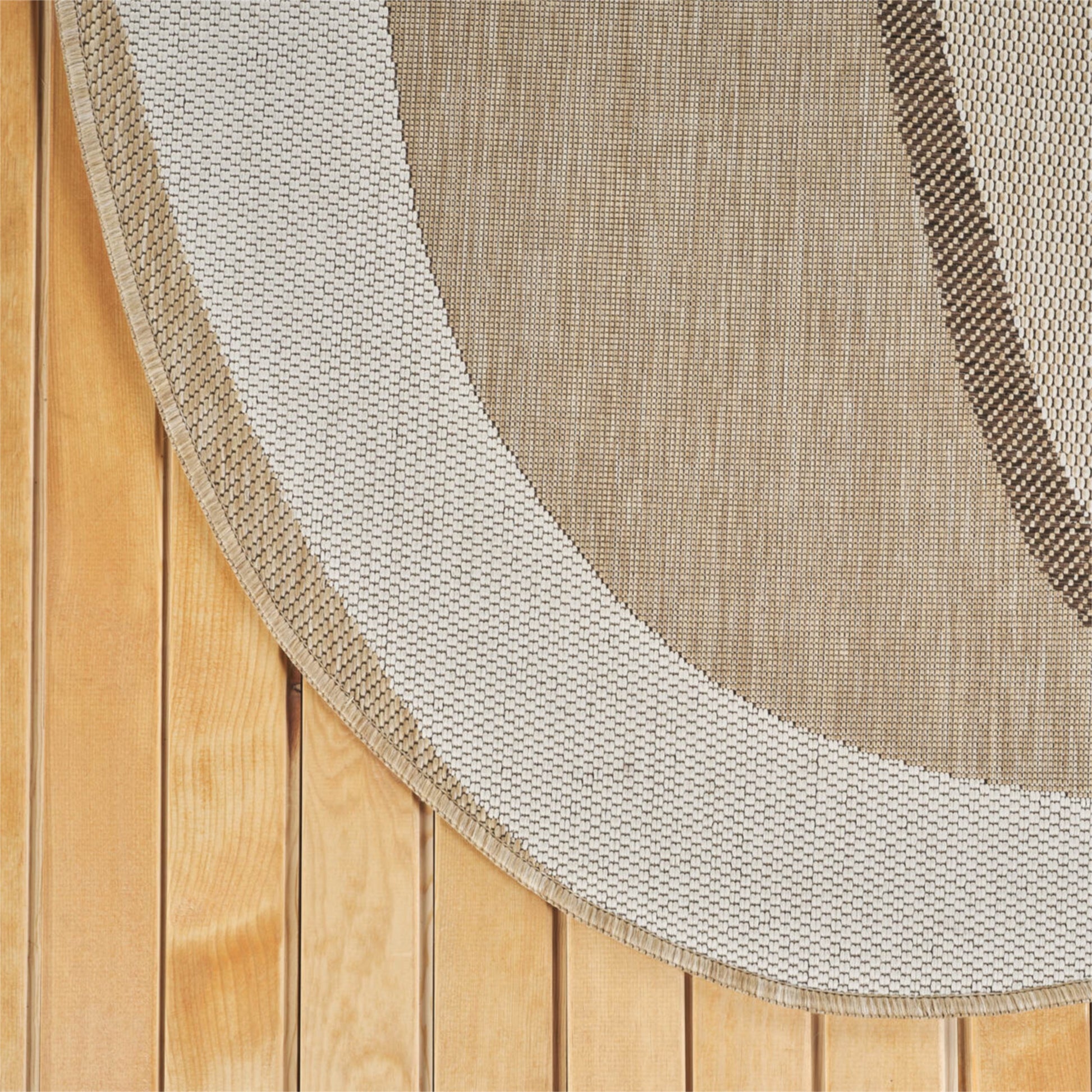 A close-up of a woven oval rug with a thick cream border and a tan center, laid over a light wood plank floor.