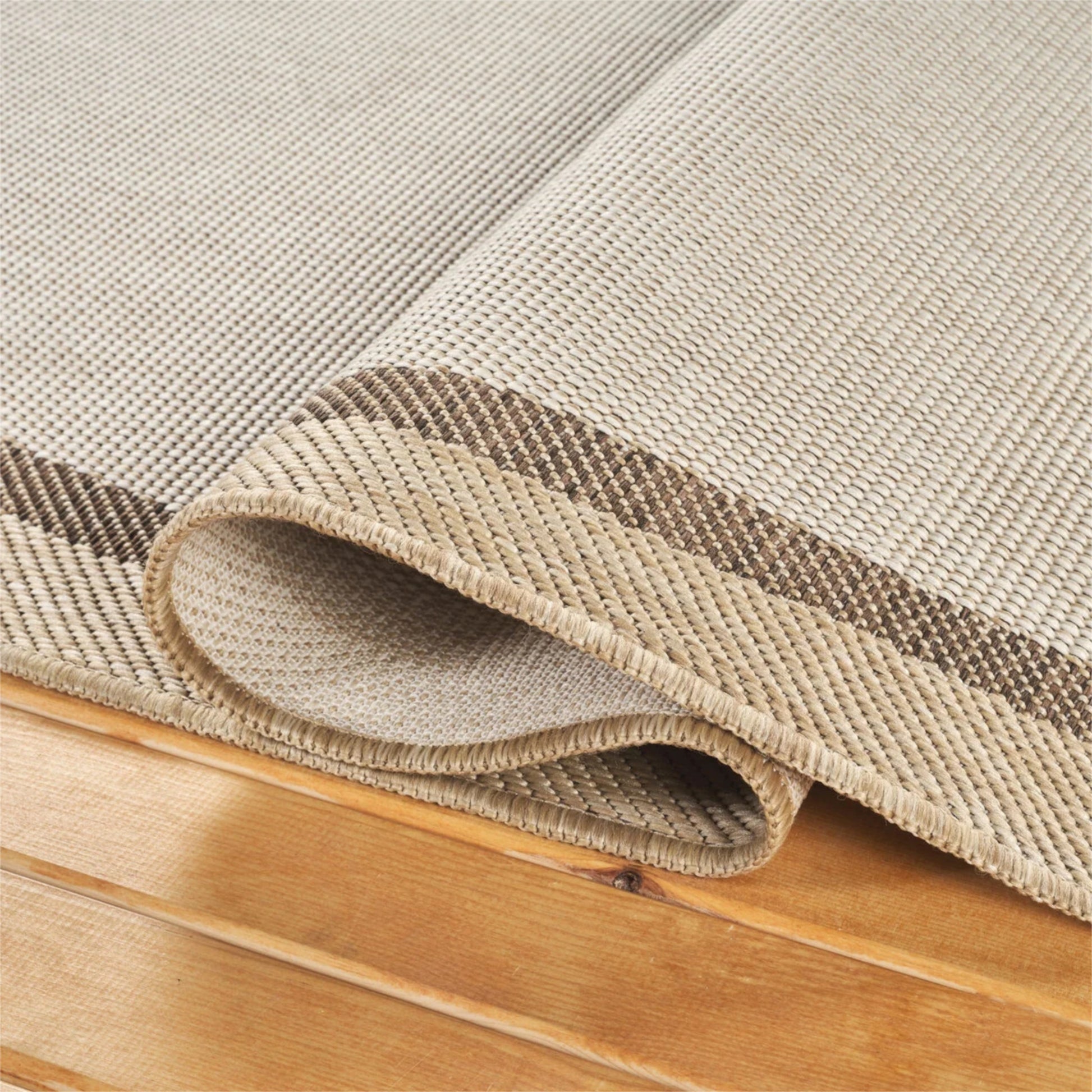 A close-up of a woven area rug in warm beige and taupe tones, shown partially rolled over to reveal its texture and underside, resting on a light wood floor.