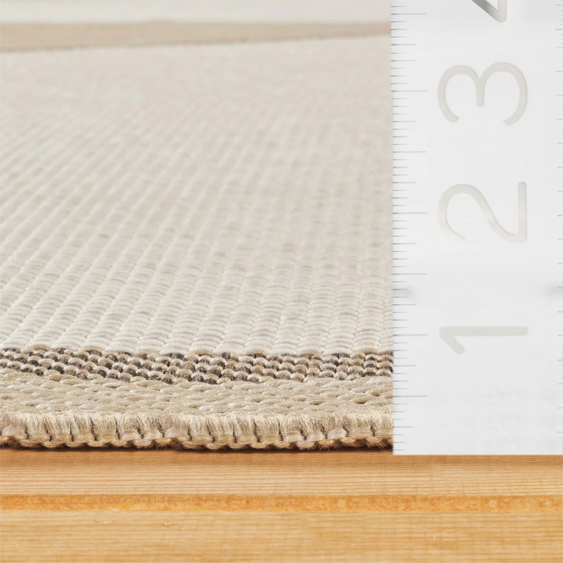 Close-up of a woven beige rug with a textured ribbed edge, shown next to a white ruler for scale on a light wood surface.