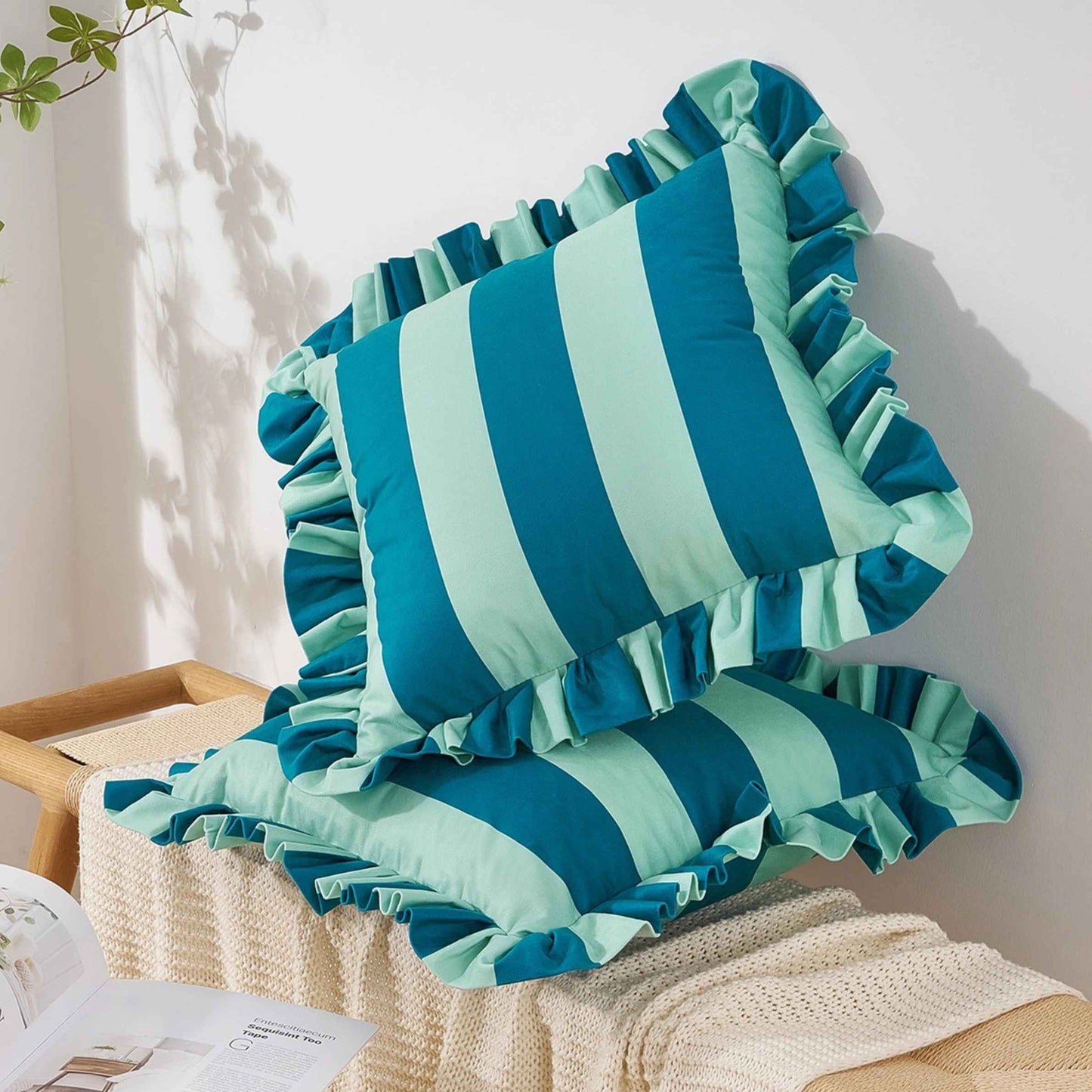 Two striped ruffled throw pillows in blue and light blue, styled on a cream knit blanket on a wooden bench against a white wall.