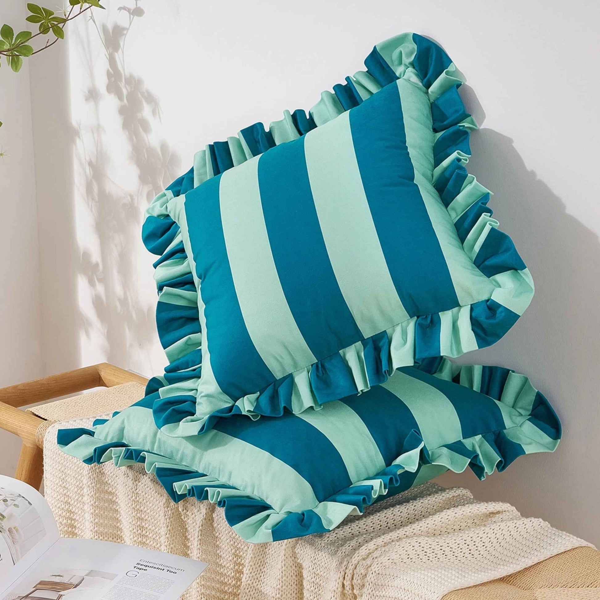 Two striped ruffled throw pillows in blue and light blue, styled on a cream knit blanket on a wooden bench against a white wall.