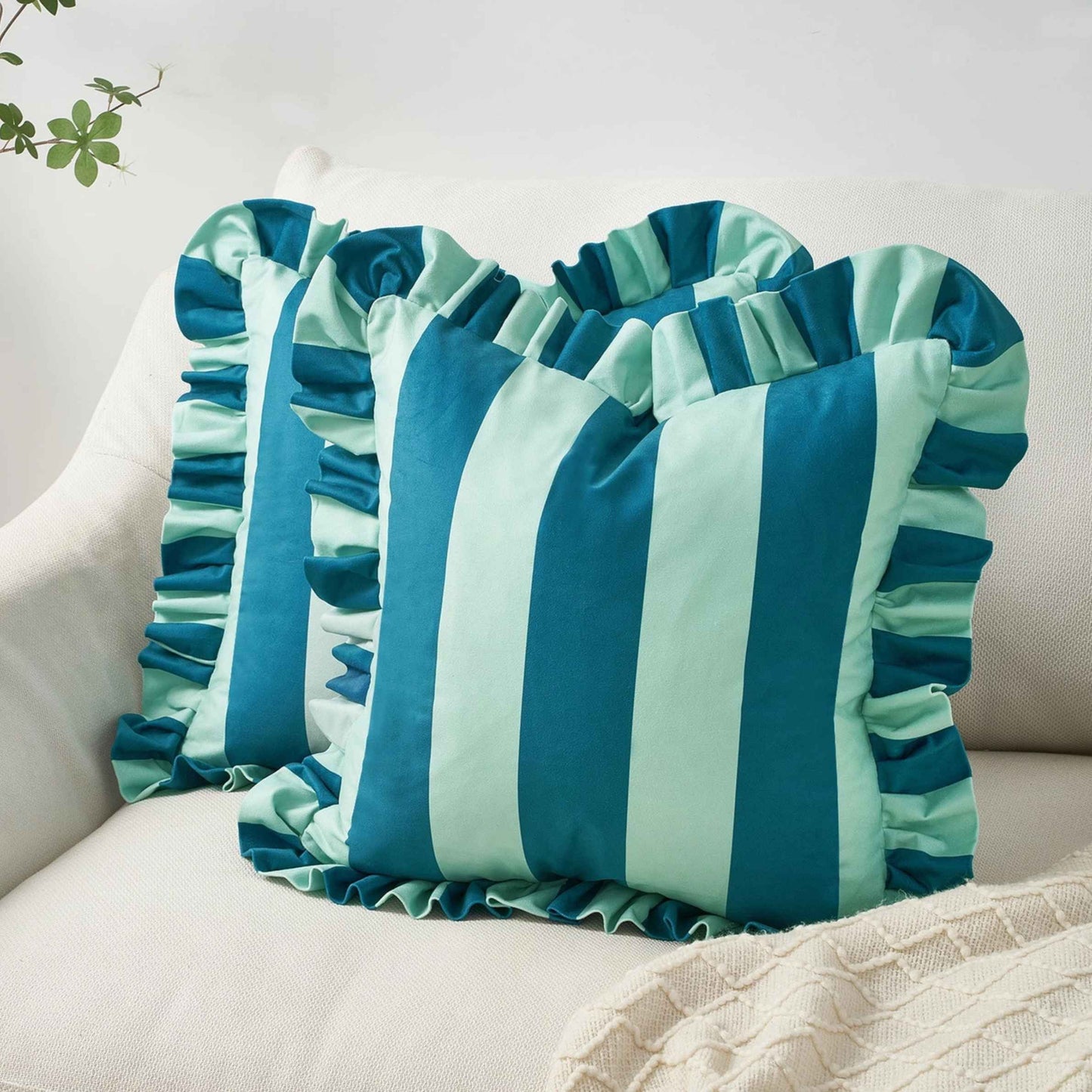 A square decorative throw pillow with vertical blue and light blue stripes and ruffled trim, styled on a cream-colored sofa.