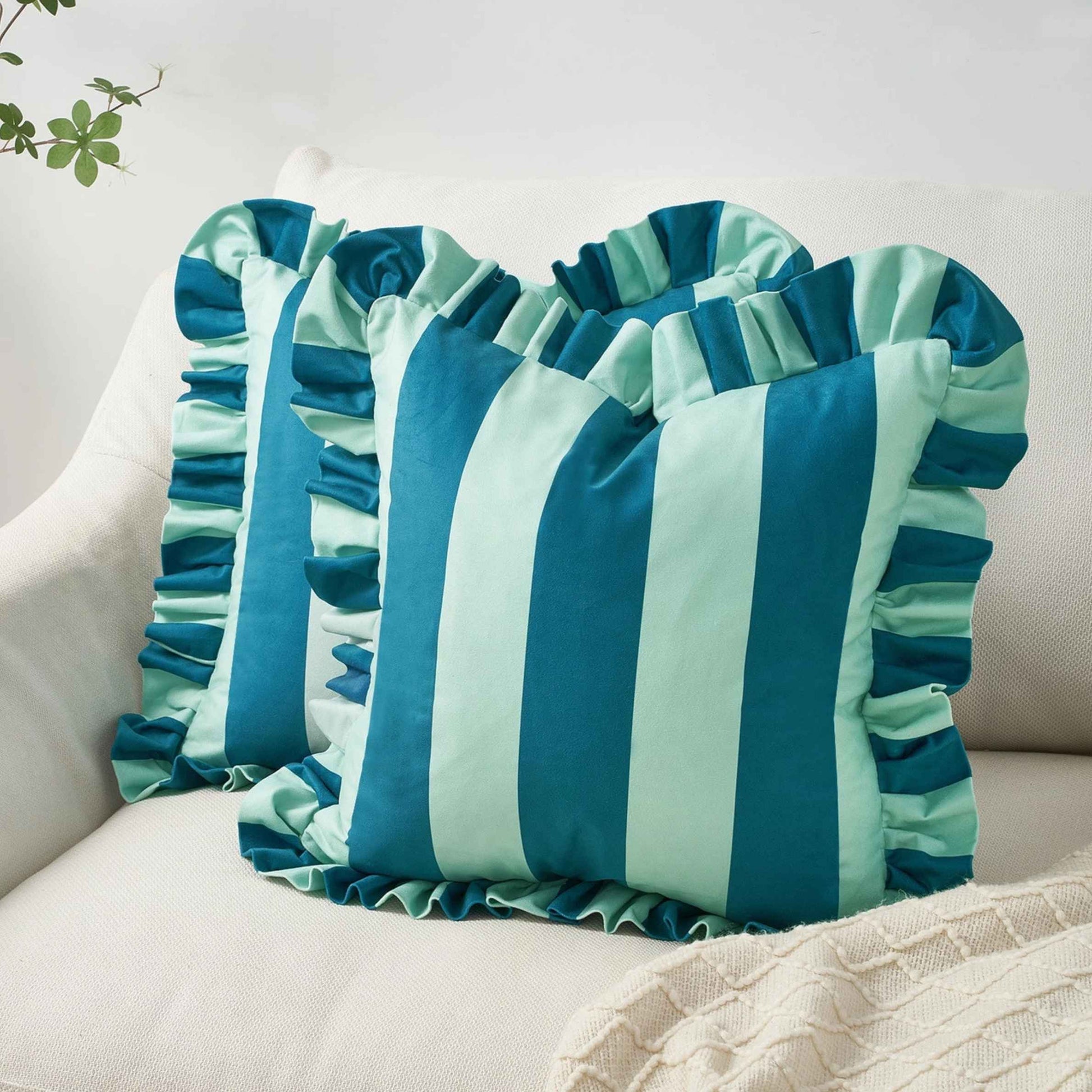 A square decorative throw pillow with vertical blue and light blue stripes and ruffled trim, styled on a cream-colored sofa.