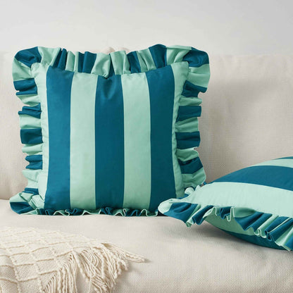 A square decorative throw pillow with vertical blue and light blue stripes and ruffled trim, styled on a cream fabric sofa next to another matching pillow.