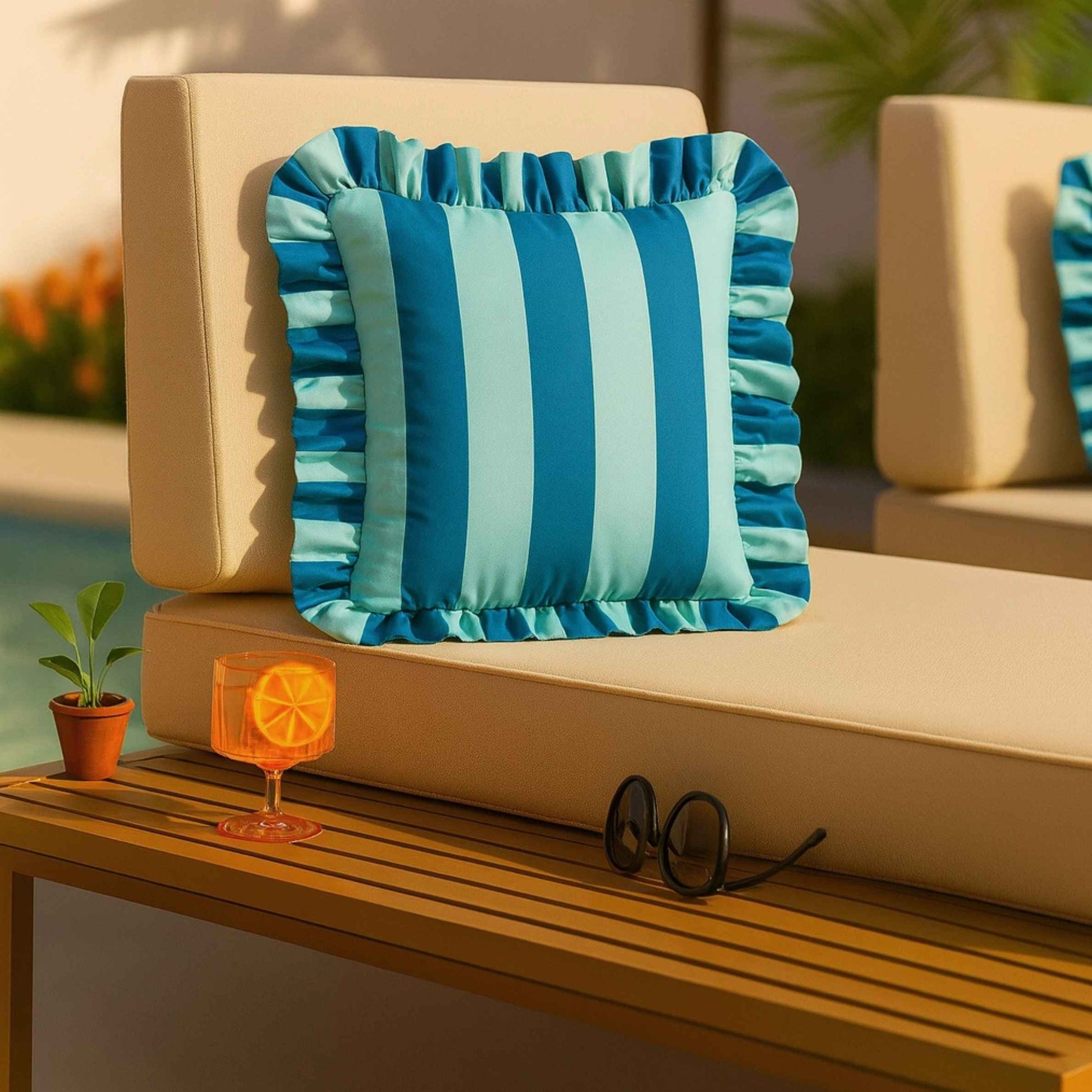 A square outdoor throw pillow with vertical blue and light blue stripes and a ruffled edge, styled on a beige patio lounge chair next to a wooden table with a drink and sunglasses.