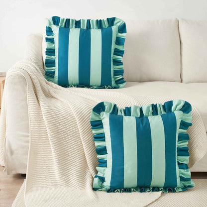 Two square throw pillows with vertical blue and light blue stripes and ruffled edges, styled on a cream-colored sofa with a textured cream blanket.