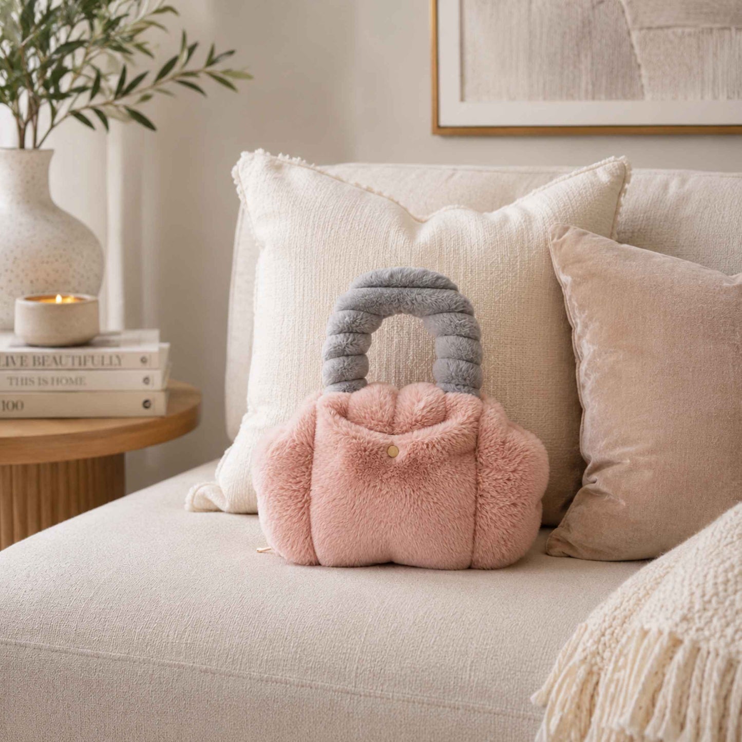 A plush, fuzzy Pur-low in dusty pink with a ribbed gray handle, styled on a cream-colored sofa with neutral-toned pillows and a side table in the background.