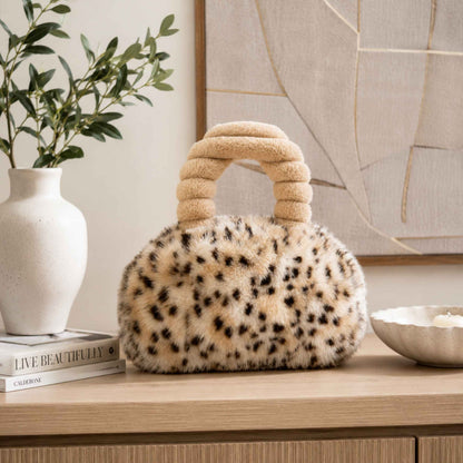 A plush leopard-print Pur-Low with a rounded body and thick ribbed handle sits on a light wood table in a styled interior setting with a white vase, book, and abstract wall art in the background.