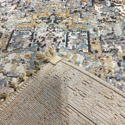 Close-up of a patterned area rug corner showing woven texture, tassels, and a beige end piece, with the rest of the rug in the background featuring a distressed multicolor design.