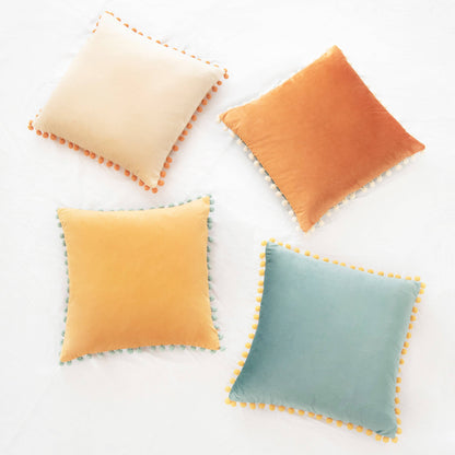 Four square pillows with pom-pom trim in beige, orange, yellow, and blue on a white background.