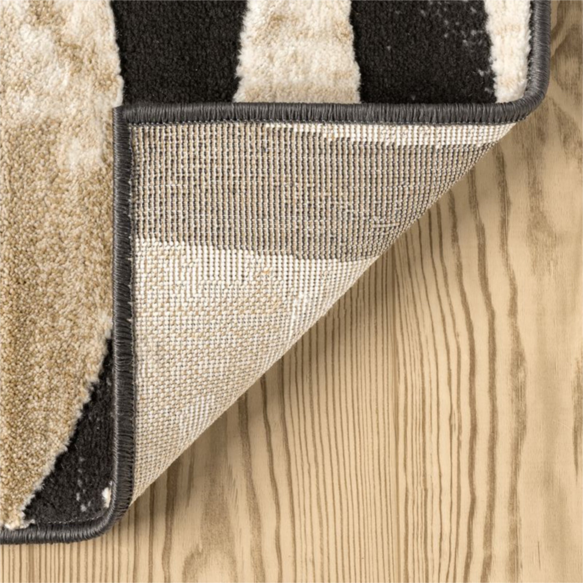 Close-up of a patterned area rug corner with beige, cream, and black stripes, showing the textured underside and dark edge trim on a light wood floor.