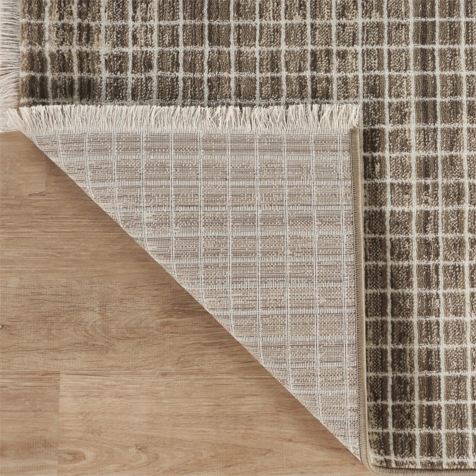 A close-up of a woven area rug with a brown and beige grid pattern, showing one corner folded over to reveal the underside and fringed edge, placed on a light wood floor.