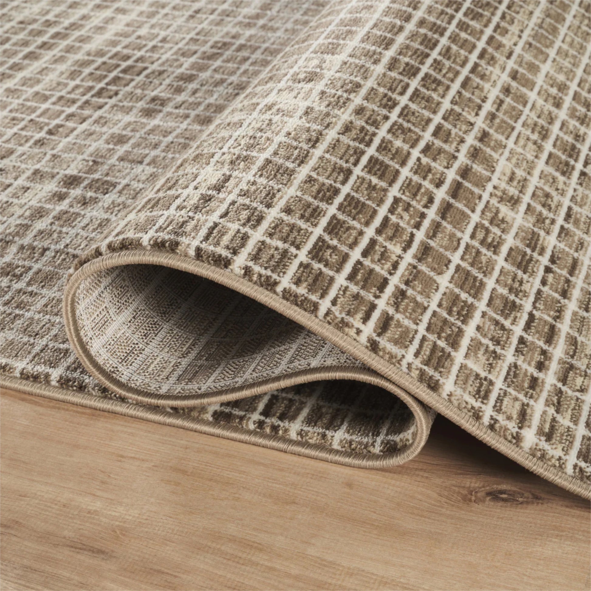 A close-up of a textured area rug with a beige and brown grid pattern, partially rolled over to show its thickness and underside, resting on a light wood floor.