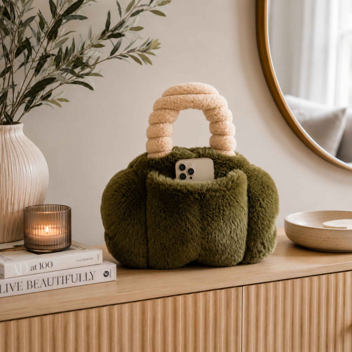 A plush moss green Pur-low with a cream handle sits on a wooden console table in a styled room setting, with a candle, books, a plant, and a mirror in the background.