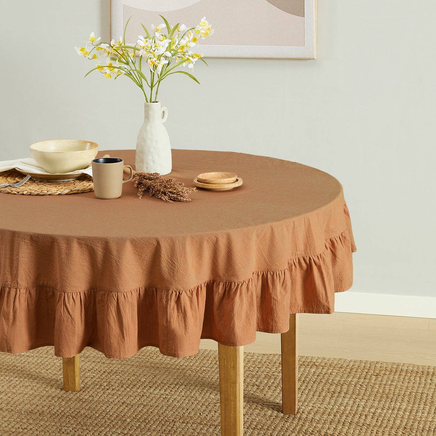 Round table with a brown ruffled tablecloth, mug, vase, and flowers in a room.