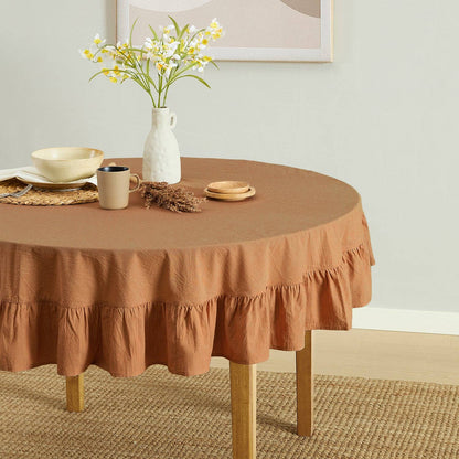 Round table with a brown ruffled tablecloth, mug, vase, and flowers in a room.