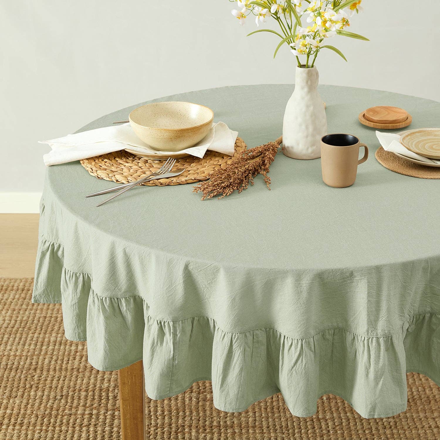 Round table with a green tablecloth, plates, cups, and a vase with flowers on a light background.