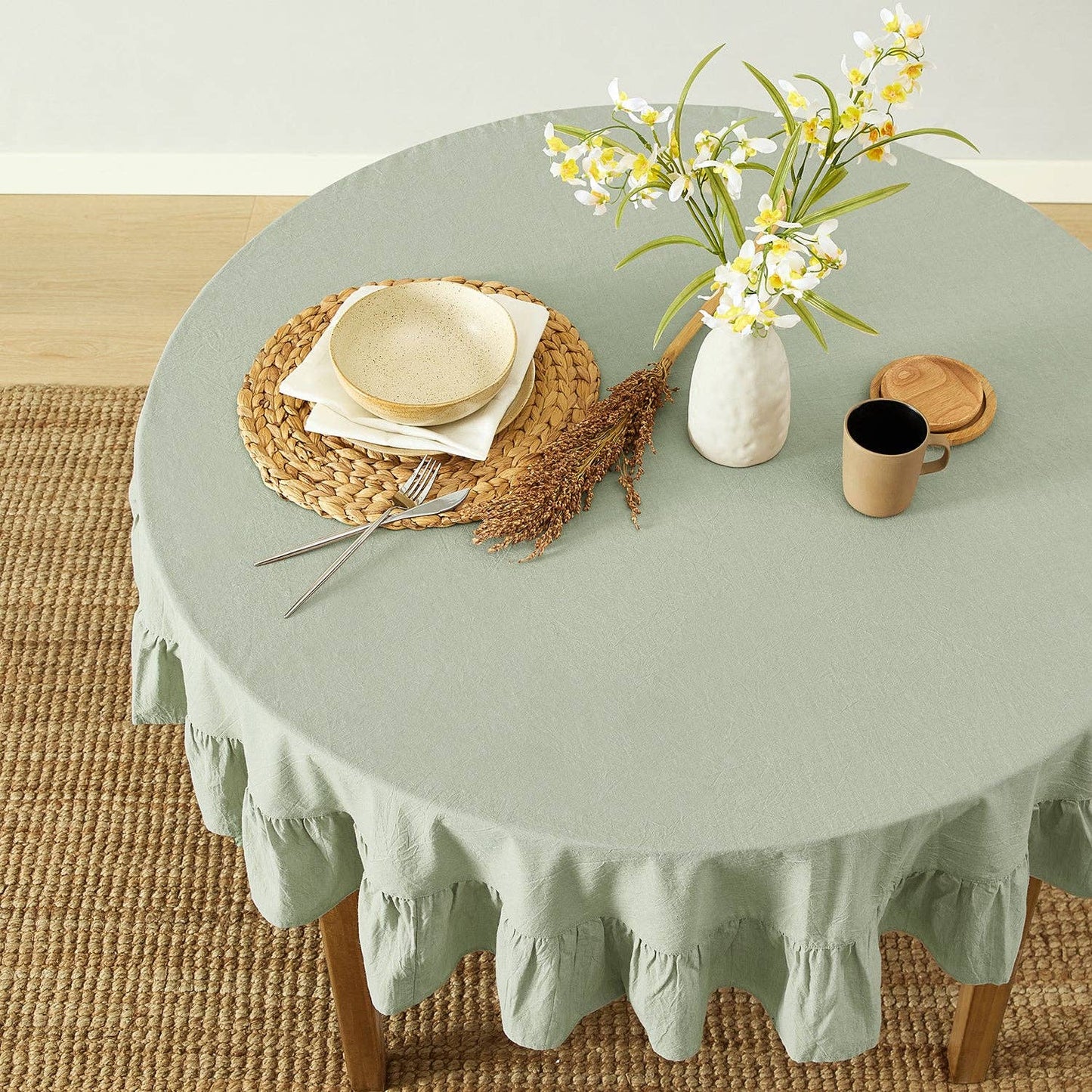 Round table with a green tablecloth, plates, cutlery, and a vase with flowers on a wooden floor.