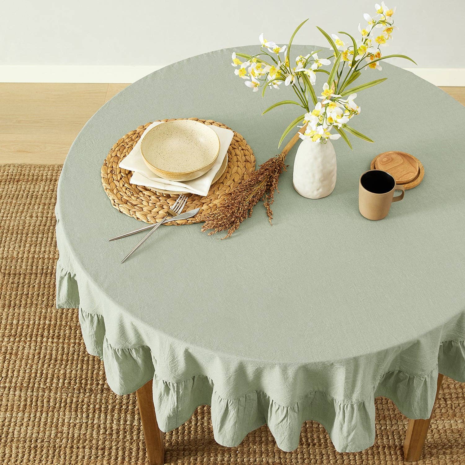 Round table with a green tablecloth, plates, cutlery, and a vase with flowers on a wooden floor.