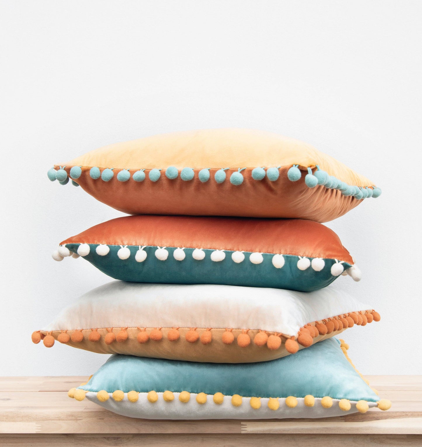 Stack of colorful pillows with pom-pom trim on a light background