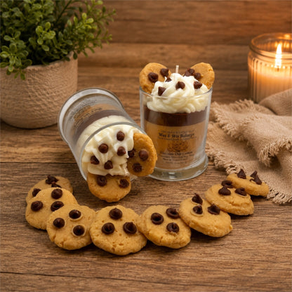 Two decorative candles styled like chocolate chip cookies in glass jars, with whipped cream and cookie pieces on top, arranged on a wooden surface with scattered cookies, a plant, and a lit candle in the background.