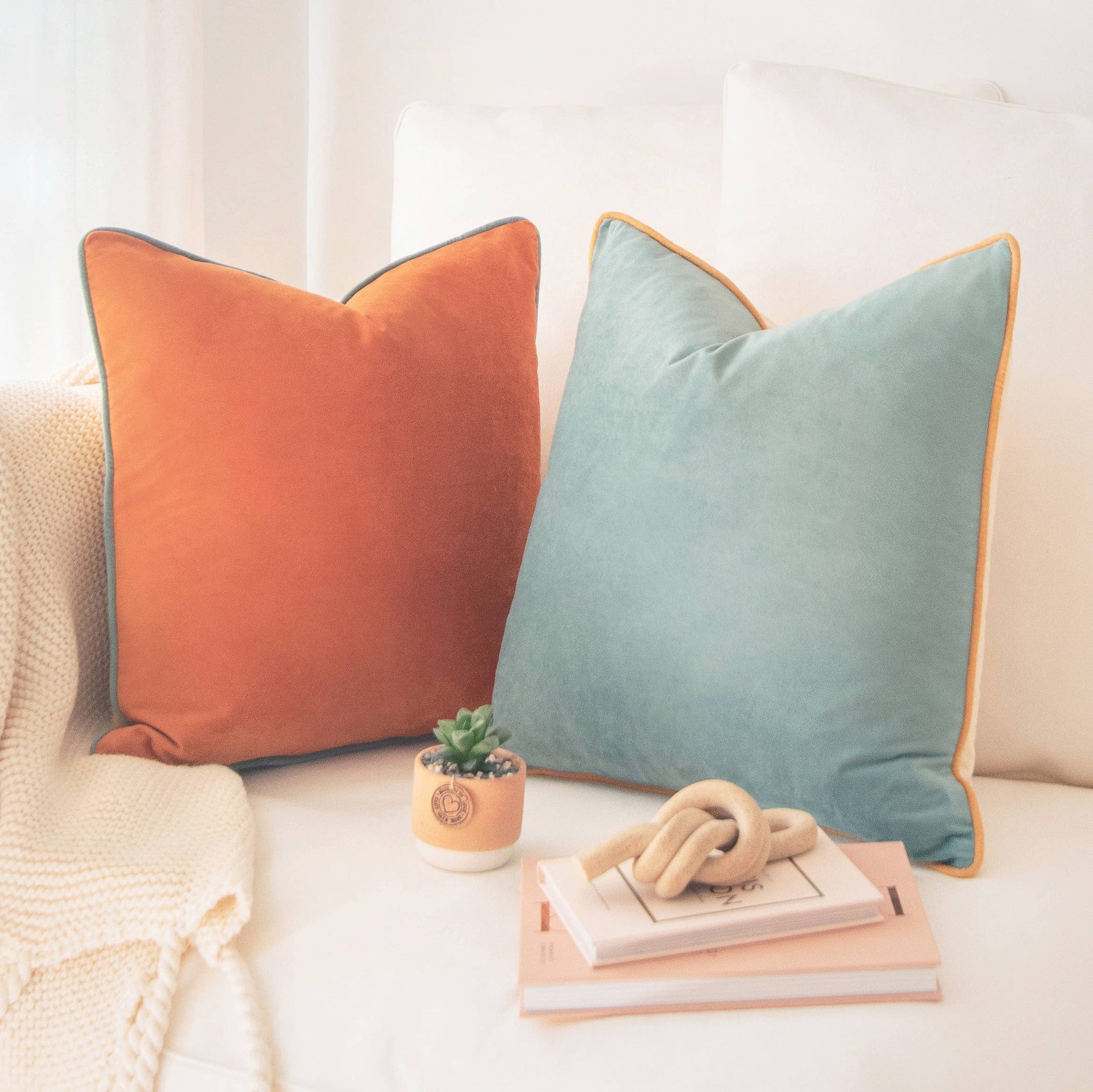 Two decorative pillows, one orange and one blue, on a sofa with a small plant and book.
