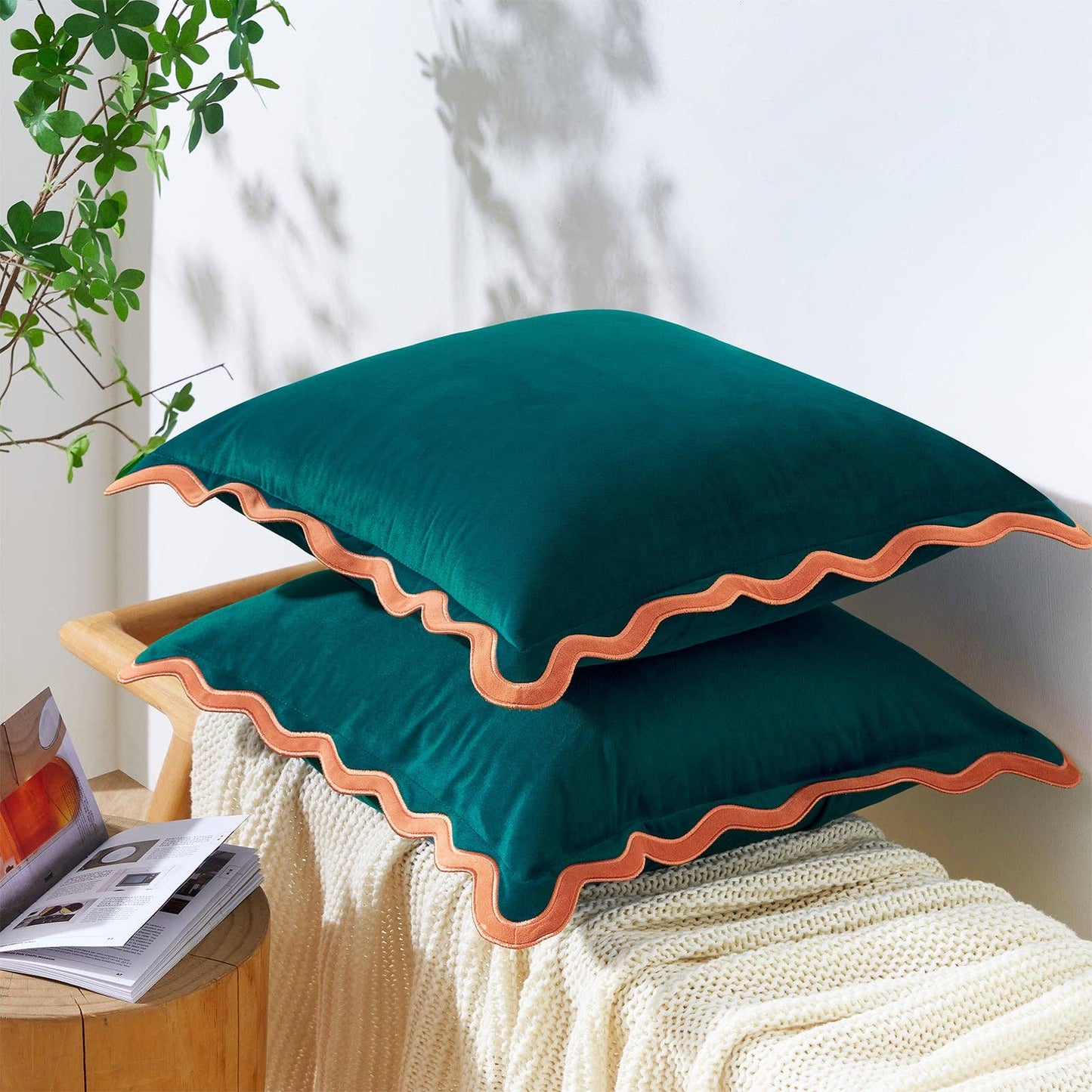 Two green pillows with orange trim on a white surface, with a plant and magazine in the background.