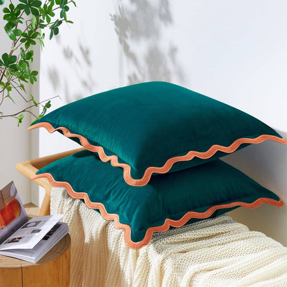 Two green pillows with orange trim on a white surface, with a plant and magazine in the background.