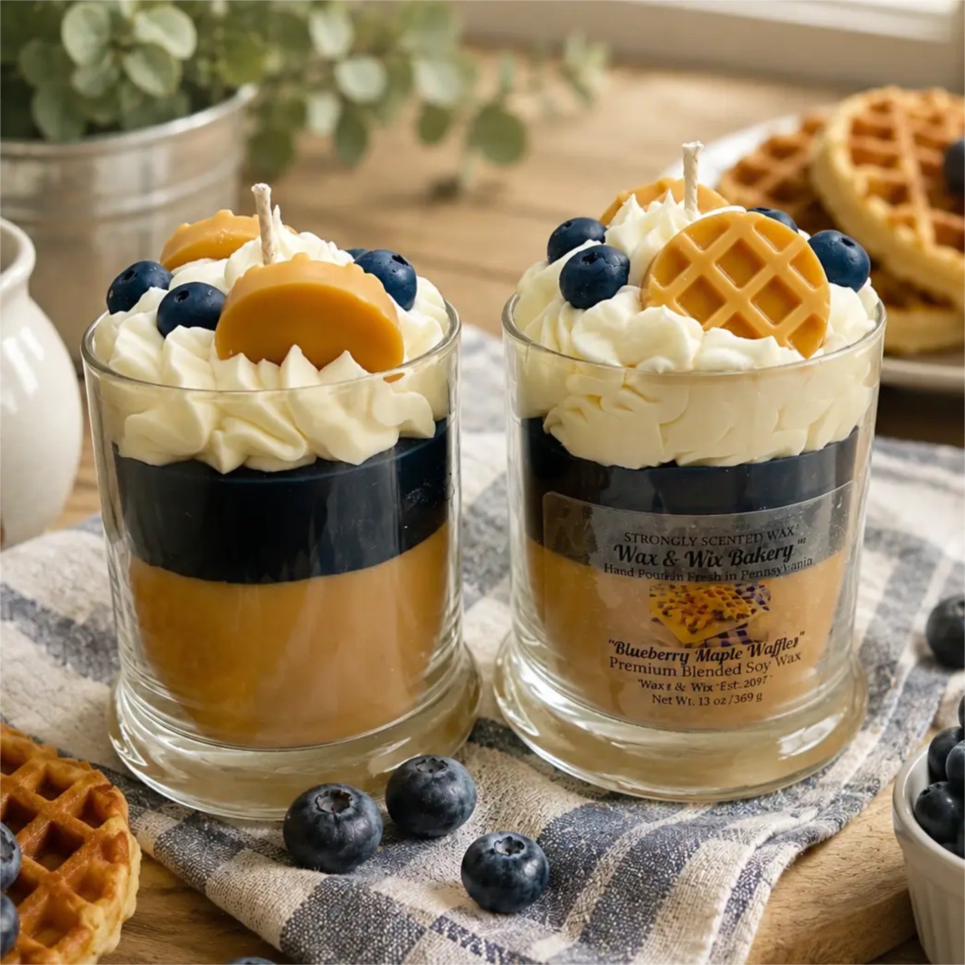Two layered glass candles styled like blueberry waffle batter, topped with whipped cream, blueberries, and waffle pieces, set on a table with waffles and a cloth. The front candle has a visible label reading 'Blueberry Maple Waffles' and 'Waffle Bakery'.
