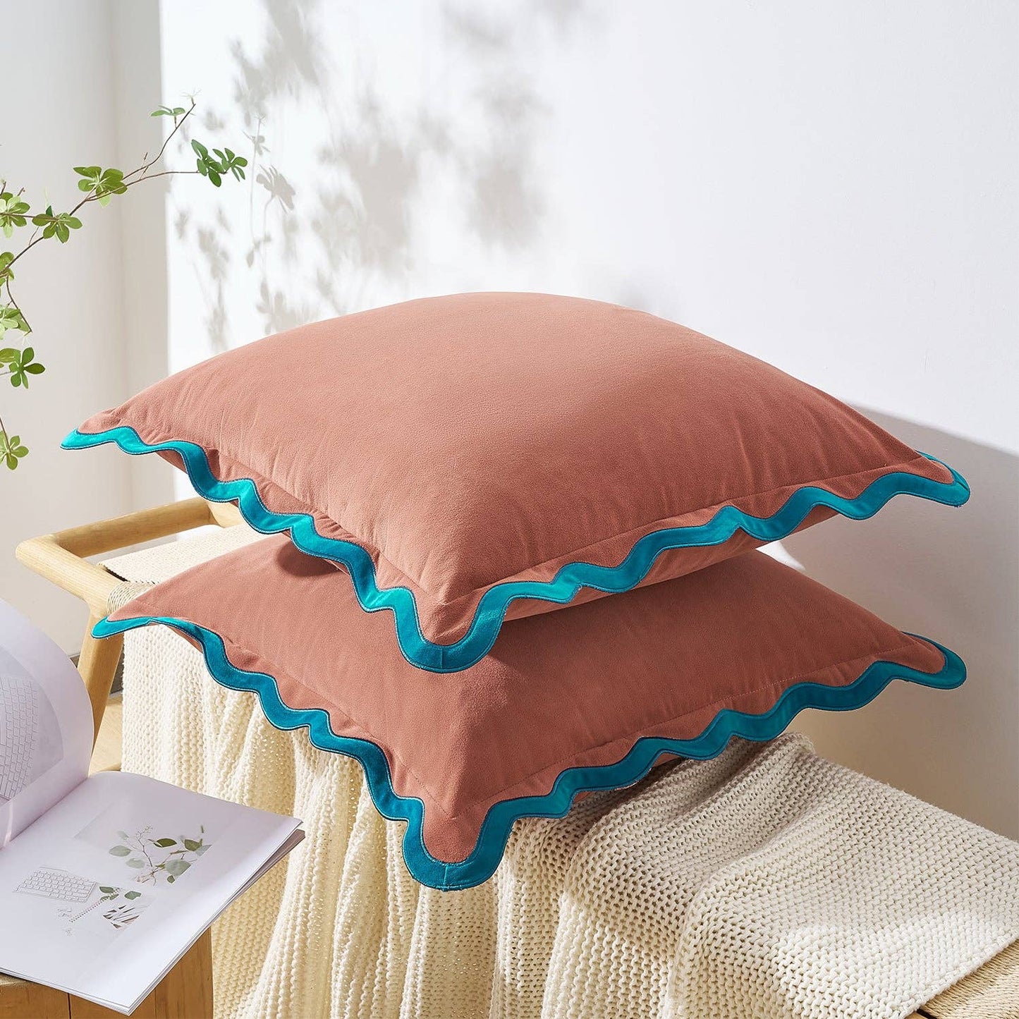 Two pink pillows with blue trim on a white surface, with a plant and book in the background.