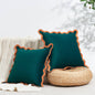 Two teal pillows with orange trim on a white surface with a plant in the background