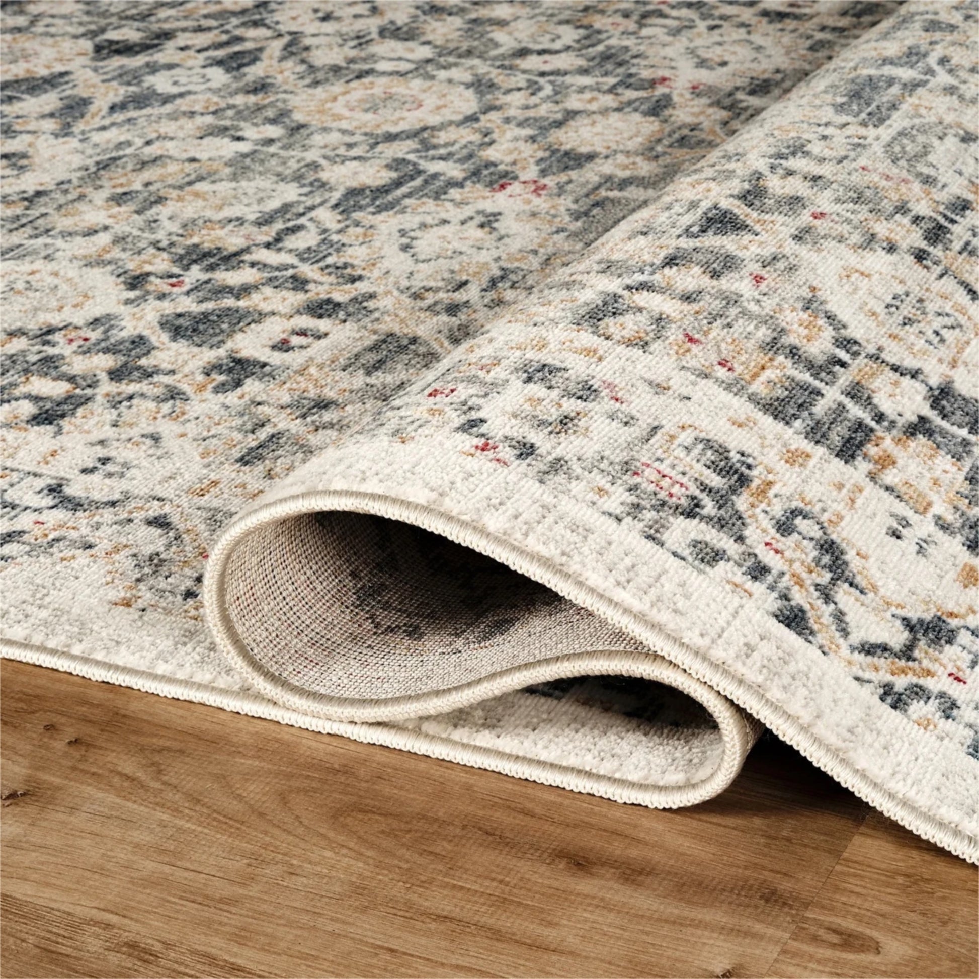 A close-up of a rolled area rug with an intricate distressed pattern in beige, gray, and muted blue, shown on a light brown wooden floor.