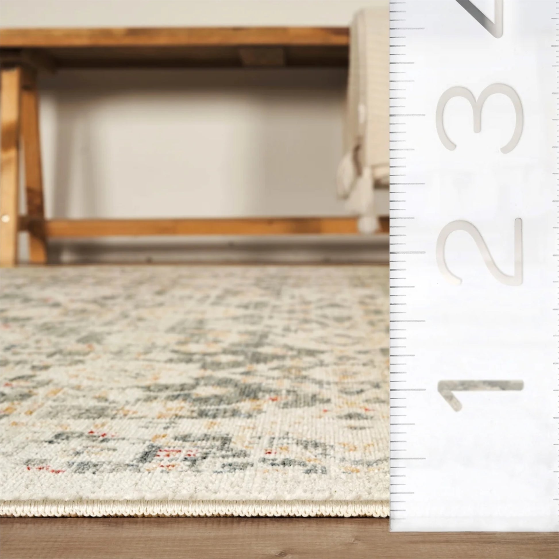 A close-up of a patterned area rug in a room setting, with a wooden bench in the background and a white ruler on the right side showing measurements.
