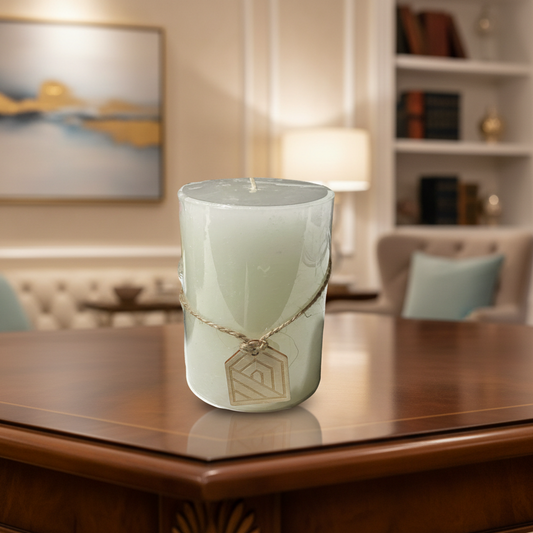 A clear pillar candle with a label and a piece of wood with a geometric design attached to it, set on a reflective surface.