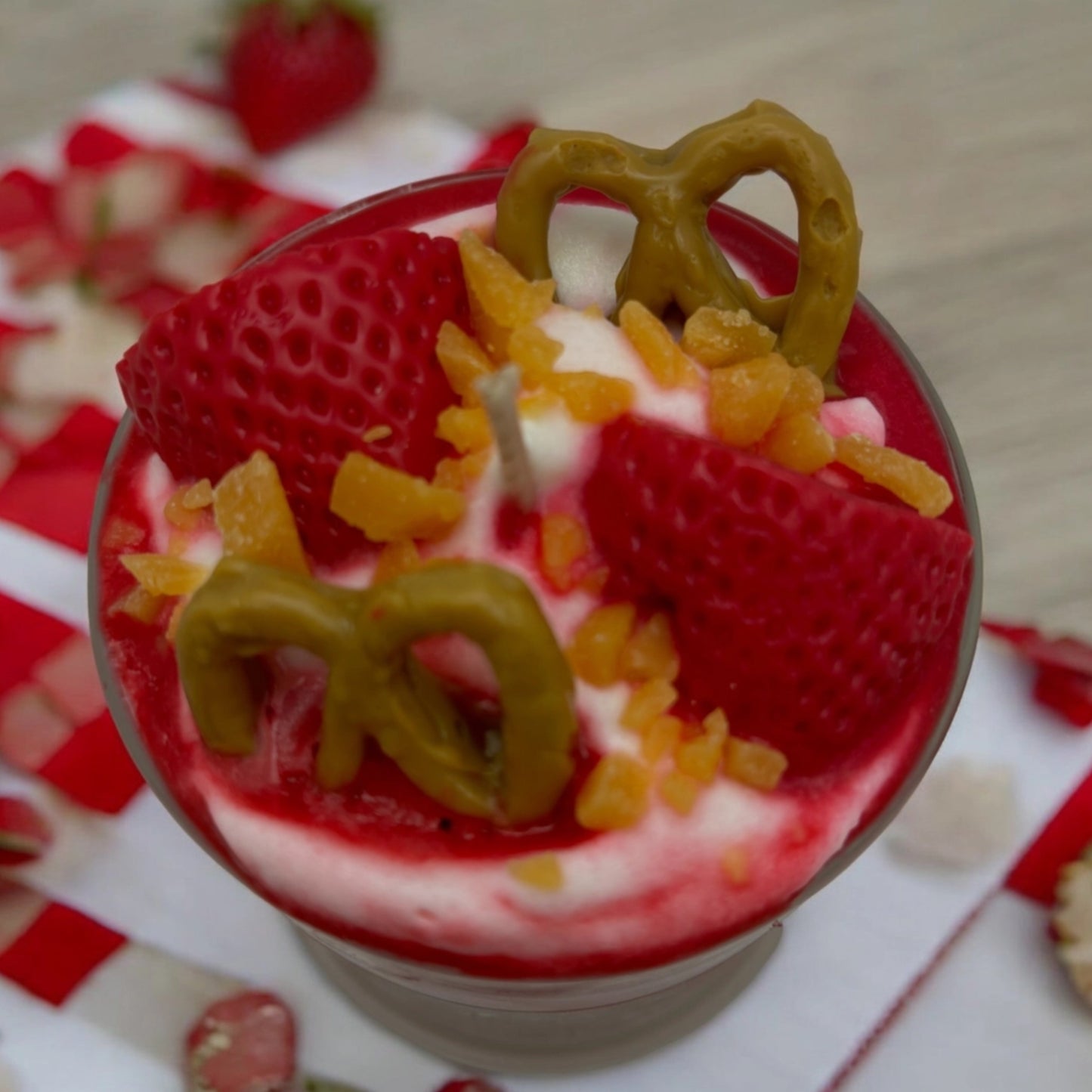 A close-up of a candle in a clear glass bowl, topped with red strawberry slices, white cream, orange candied fruit pieces, and pretzel-shaped decorations on a red-and-white checkered cloth.