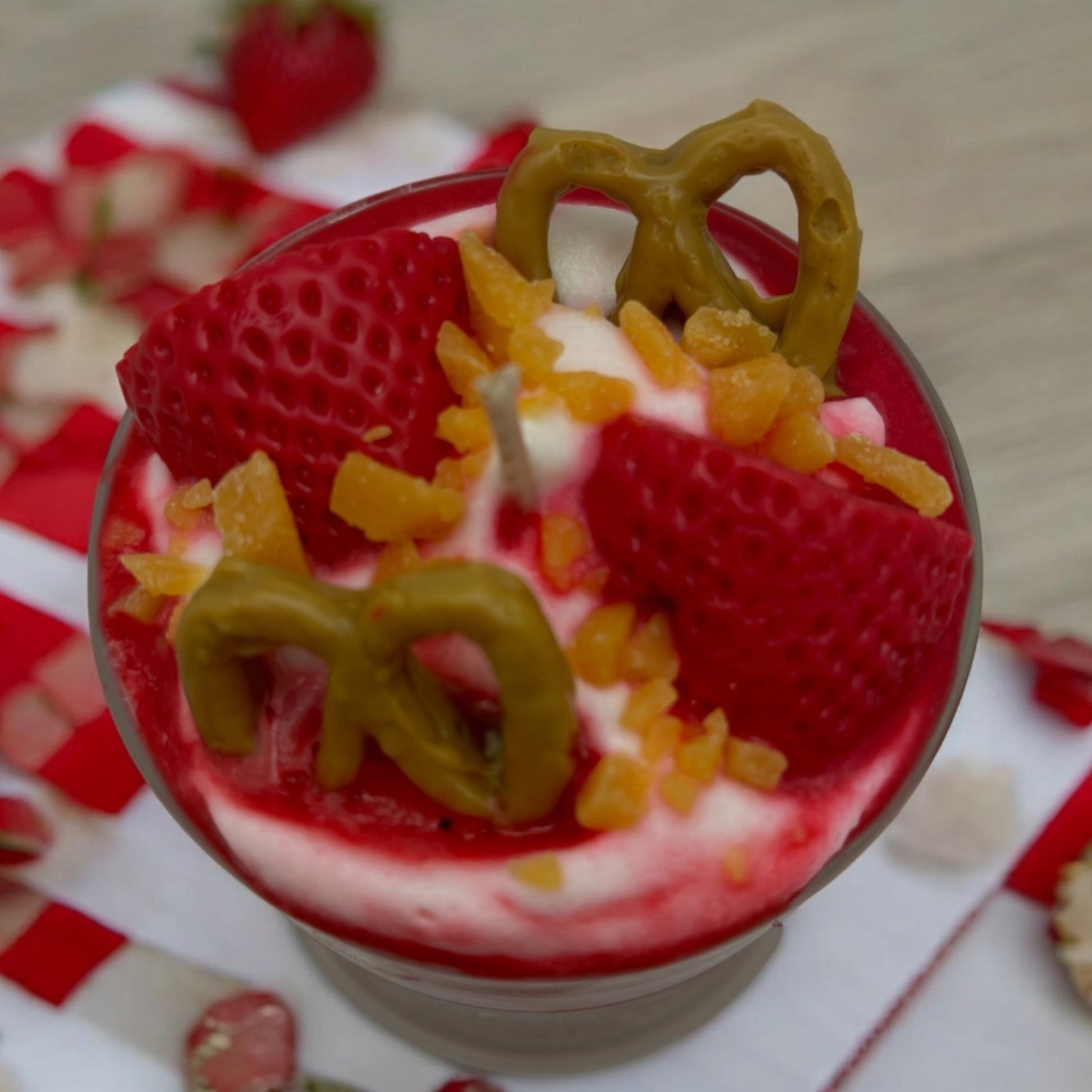 A close-up of a candle in a clear glass bowl, topped with red strawberry slices, white cream, orange candied fruit pieces, and pretzel-shaped decorations on a red-and-white checkered cloth.
