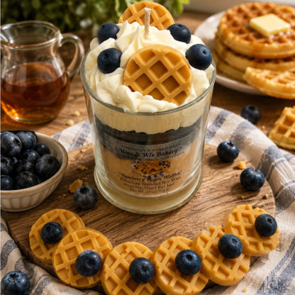 A decorative candle in a clear glass jar styled like a waffle-pan breakfast dessert, filled with cream-colored wax and topped with whipped cream, blueberries, and a waffle piece. The candle sits on a wooden board surrounded by waffles and blueberries, with a glass of syrup and stacked waffles in the background.