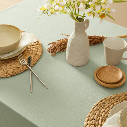 Dining table setting with ceramic plates, cutlery, a vase with flowers, and wooden coasters on a light green tablecloth.