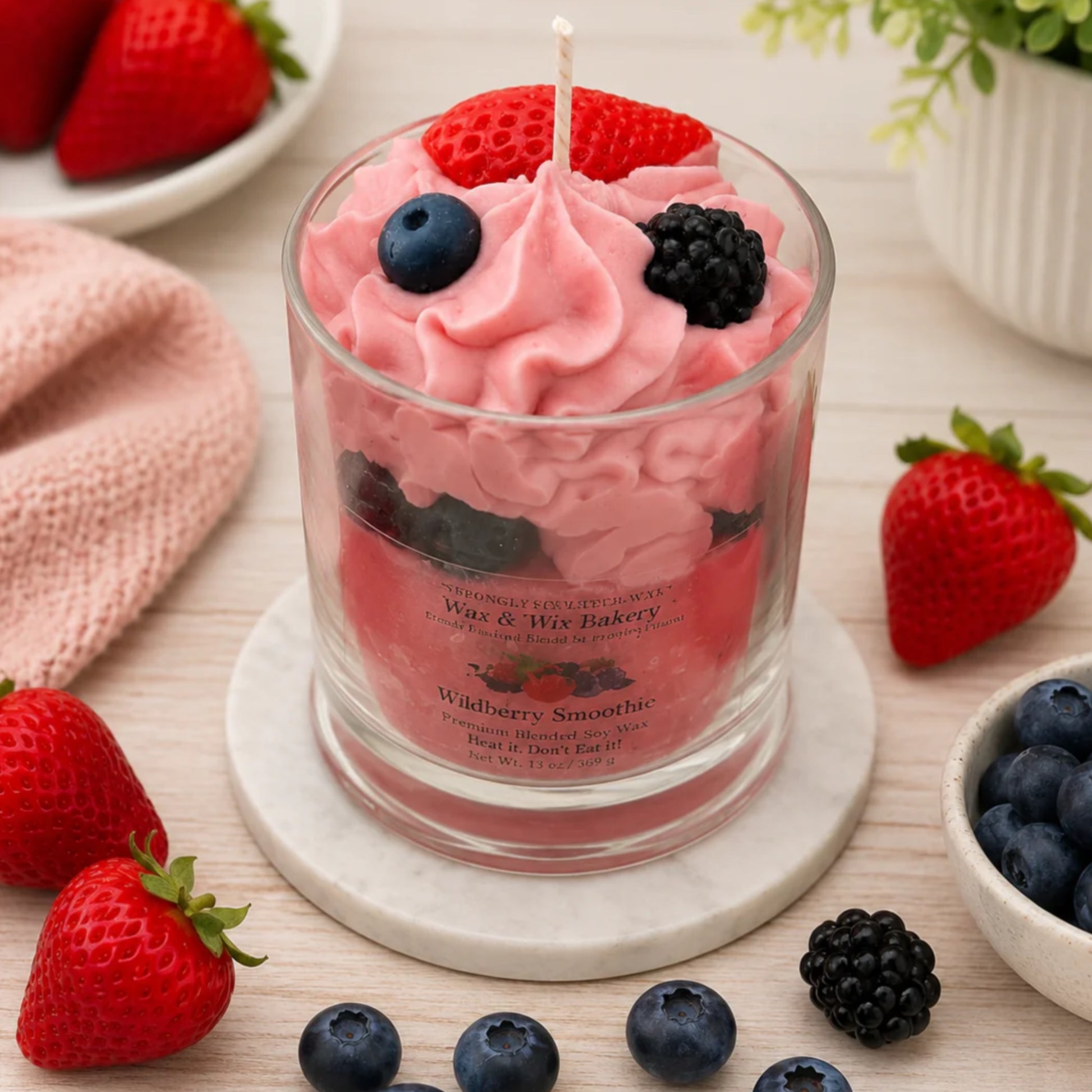 A fruit-flavored candle in a clear glass jar with a pink whipped-cream style wax top and visible wick, styled on a marble coaster with strawberries, blueberries, and a pink cloth around it. The label reads Wildberry Smoothie and is branded Wase & Mrs.